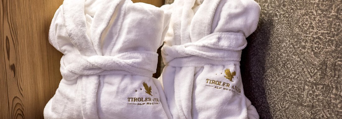 Two white bathrobes are lying on a cozy sofa. They are adorned with a logo and radiate a relaxed atmosphere. - Alp Resort Tiroler Adler - Sölden