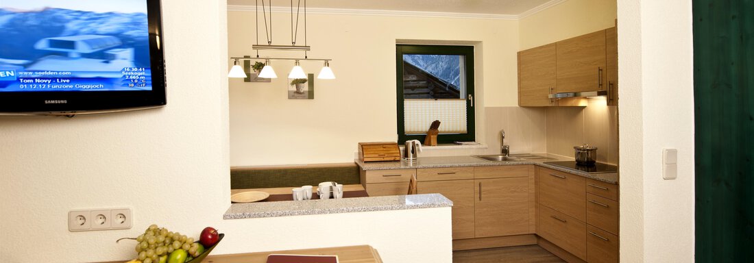 A modern kitchen with wooden shelves and cooking appliances. In the background, there is a television and a window with a view outside. - Alp Resort Tiroler Adler - Sölden