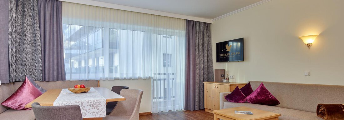 A modern living room with wood-colored flooring and a large window. The space is furnished with a dining table, cozy chairs, and sofas. - Alp Resort Tiroler Adler - Sölden