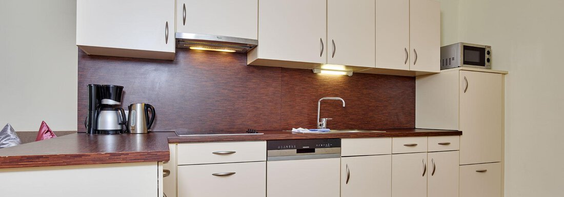 A modern kitchen with light cabinets and a wooden workspace. The equipment includes a kettle, a dishwasher, and a microwave. - Alp Resort Tiroler Adler - Sölden