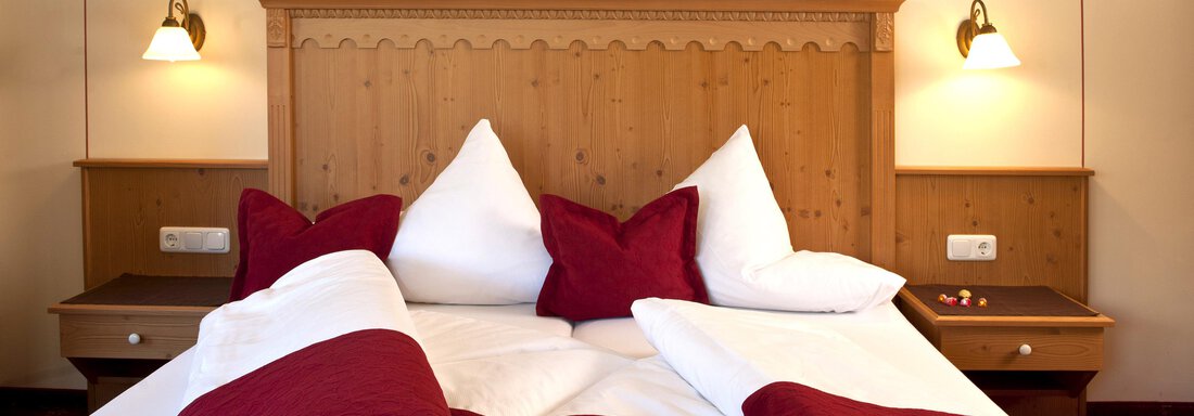 A cozy bedroom with a wooden bed and red blankets. The walls are light, and bedside lamps provide a warm light. - Alp Resort Tiroler Adler - Sölden