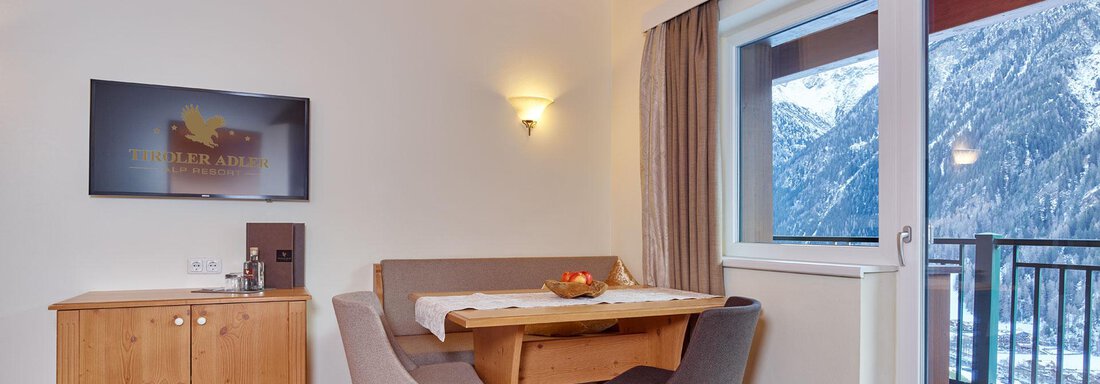 A cozy room with wooden ceilings, a dining table, and a television. Large windows offer a view of the mountains. - Alp Resort Tiroler Adler - Sölden