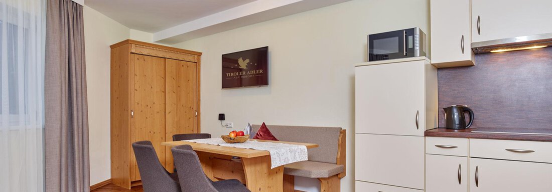 A bright, modern living room with a dining table, chairs, and a kitchenette. The decor is simple and functional, with wooden flooring and a large window. - Alp Resort Tiroler Adler - Sölden