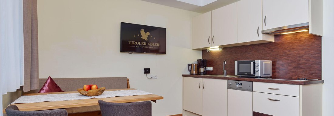 A modern kitchen with wooden flooring and a bright interior. A dining table with gray chairs and a bowl of fruit is also visible. - Alp Resort Tiroler Adler - Sölden