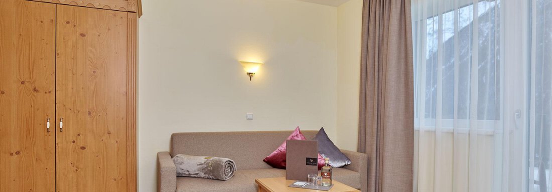 A modern living room with a beige sofa and a small wooden table. Light curtains hang in front of the window, and a cozy atmosphere invites relaxation. - Alp Resort Tiroler Adler - Sölden