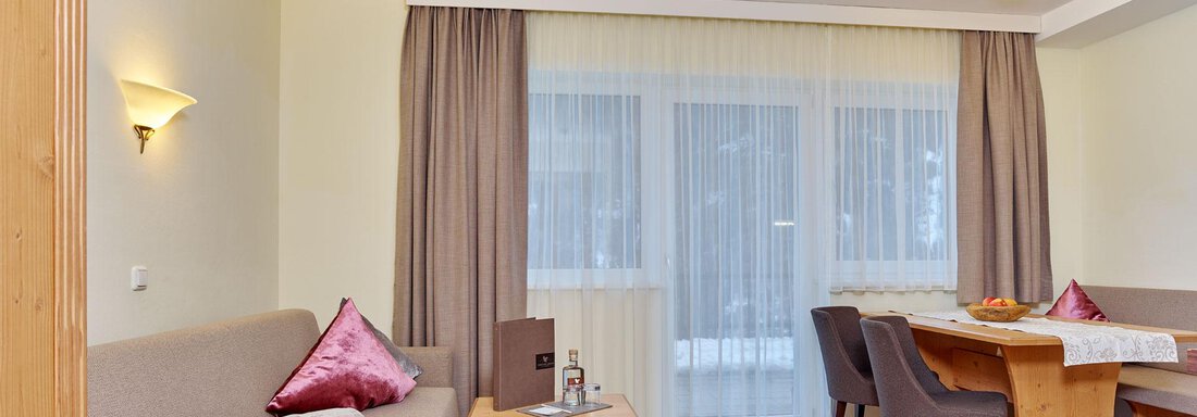 A cozy living room with a beige sofa and a wooden table. Large windows let in plenty of light, and the overall ambiance feels friendly and inviting. - Alp Resort Tiroler Adler - Sölden