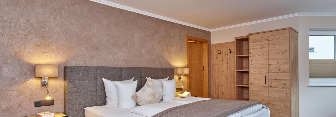 A modern bedroom with a large bed, two pillows, and a stylish duvet. The walls are painted in a soft brown tone, and there is a wooden wardrobe as well as matching nightstands. - Alp Resort Tiroler Adler - Sölden