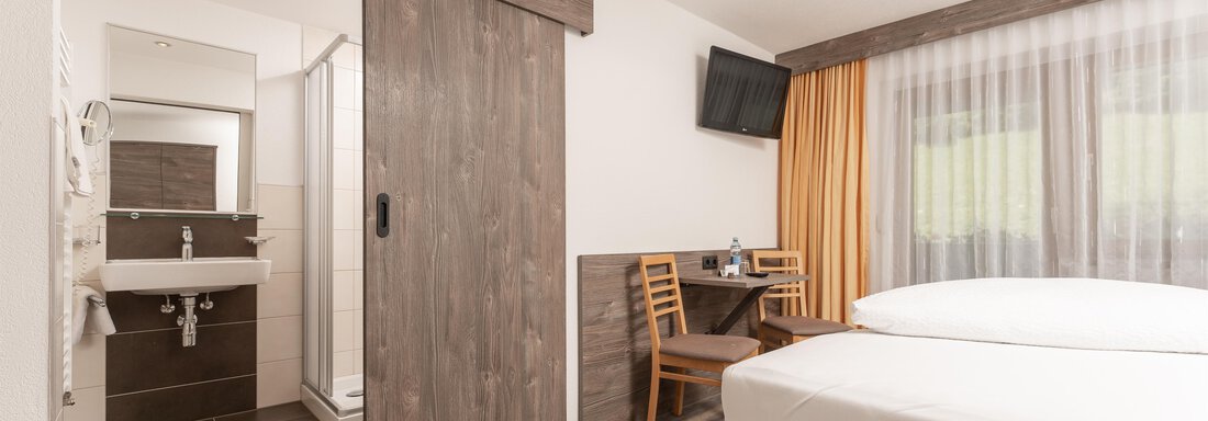 A modern hotel room with two beds and an adjoining bathroom. There are wooden details, a television, and a small living area with a table and chairs. - Alpenliebe - Sölden