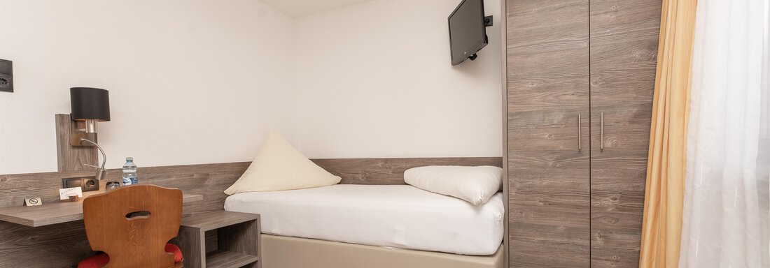 Zimmerfotos-5 A simple hotel room with a single bed, a desk, and a television. The room has bright walls and a modern wooden floor. - Alpenliebe - Sölden
