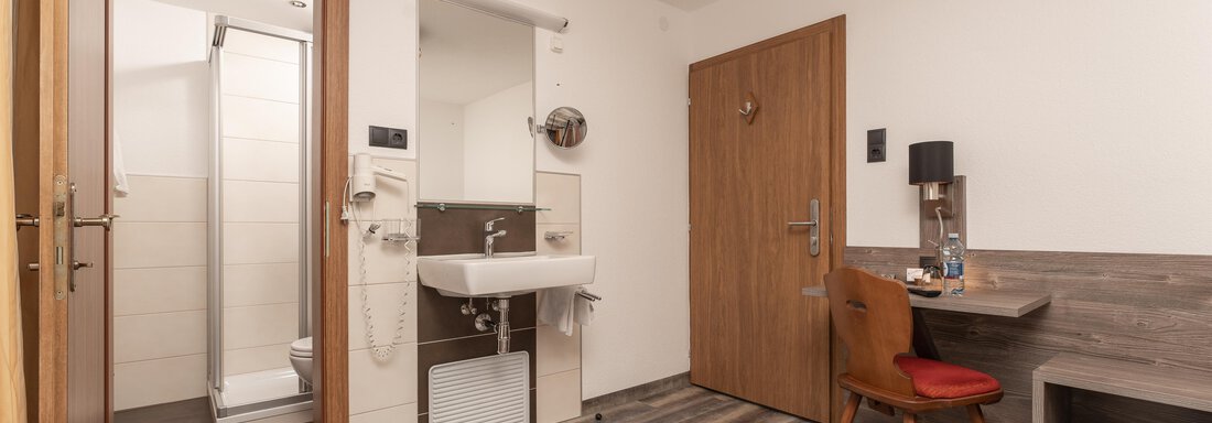 A modern hotel room with a bathroom, a desk, and a chair. The room has bright walls and a cozy wooden floor. - Alpenliebe - Sölden