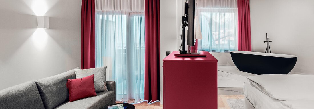 A modern hotel room with a gray sofa, a red TV table, and large windows. In the background, there is a modern bed and a freestanding bathtub. - AlpenParks The Secret Sölden - Sölden