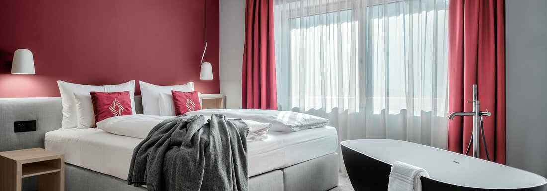 A modern bedroom with a large bed and a stylish bathtub. The walls are painted red and the windows are adorned with light curtains. - AlpenParks The Secret Sölden - Sölden