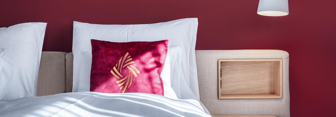 A modern bedroom with red wall paneling and a cozy bed. On the nightstand, there is a lamp and a decorative pillow. - AlpenParks The Secret Sölden - Sölden