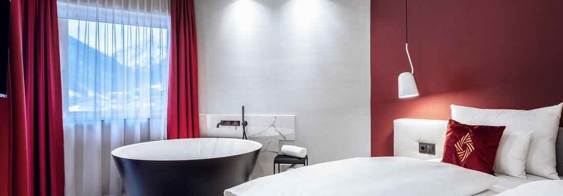 A modern hotel room with a large bed and an elegant bathroom. The walls are partly red and there is plenty of natural light from the window. - AlpenParks The Secret Sölden - Sölden