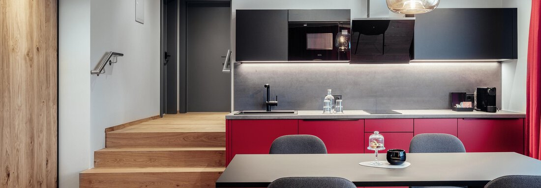 A modern kitchen with red kitchen cabinets and a cozy dining table group. The stairs lead to another room in the background. - AlpenParks The Secret Sölden - Sölden