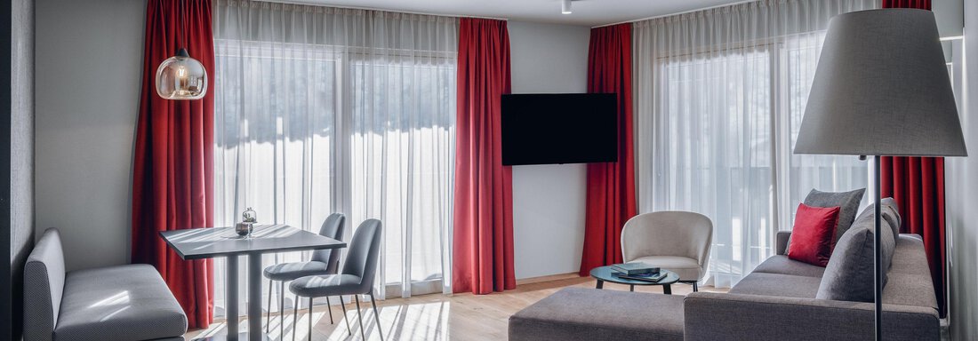 A modern living room with large windows and a red curtain. The furnishings consist of a comfortable sofa, a table, and chairs. - AlpenParks The Secret Sölden - Sölden