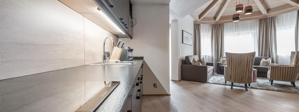 A modern kitchen with an elegant design and wooden surfaces. In the background, there is a comfortable living room with cozy seating and large windows. - AlpenParks The Secret Sölden - Sölden