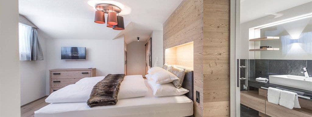 A modern bedroom with a large bed and warm wooden paneling. There is a television and an adjacent bathroom with stylish fixtures. - AlpenParks The Secret Sölden - Sölden