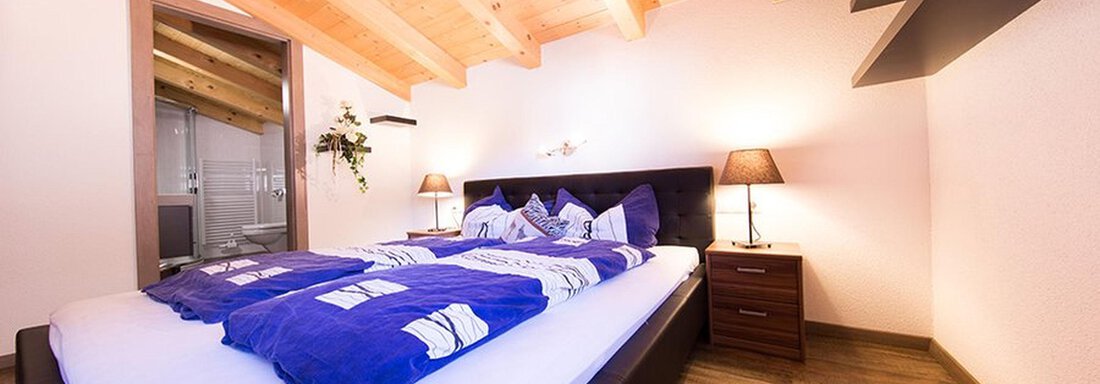 A cozy bedroom with a large bed and blue bedding. The wooden ceiling and warm light create an inviting atmosphere. - Alpensweet - Sölden