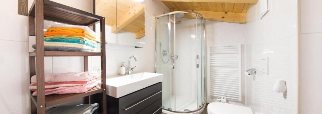 A modern bathroom with a glass shower and a white sink. There is a shelf with colorful towels and wooden ceiling surfaces. - Alpensweet - Sölden