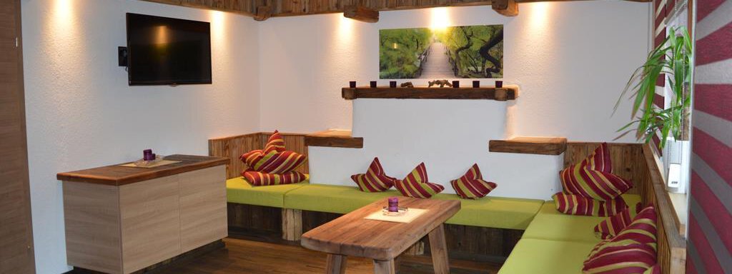 Wohnbereich A cozy living space with wooden walls and a large table. The seating area is equipped with colorful cushions and there is a television on the wall. - Am Trogwohl - Sölden