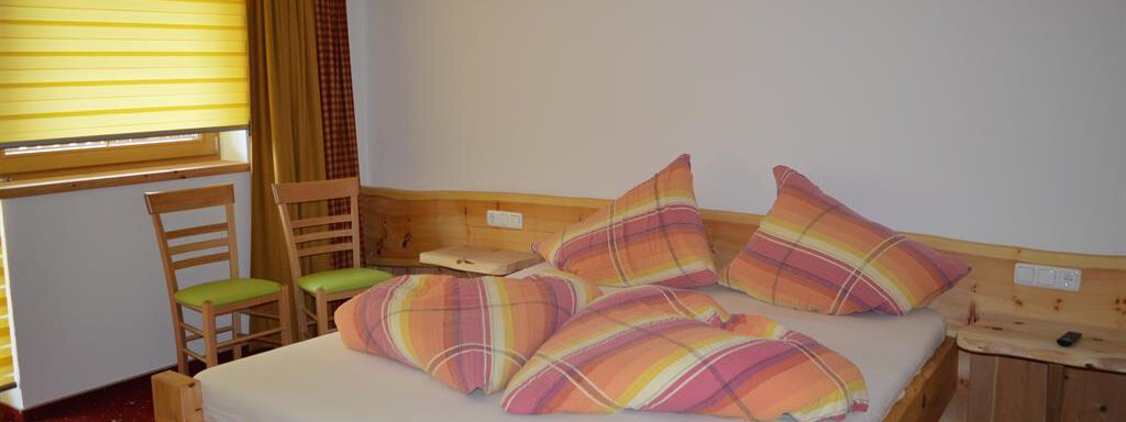 A cozy bedroom with a wood-paneled bed and colorful cushions. The windows are covered with yellow curtains, and there is also a small table with chairs. - Am Trogwohl - Sölden