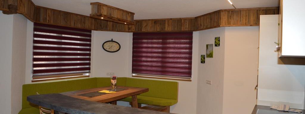 A modern kitchen with a cozy seating area in shades of green. The walls are decorated with wood paneling and red blinds. - Am Trogwohl - Sölden