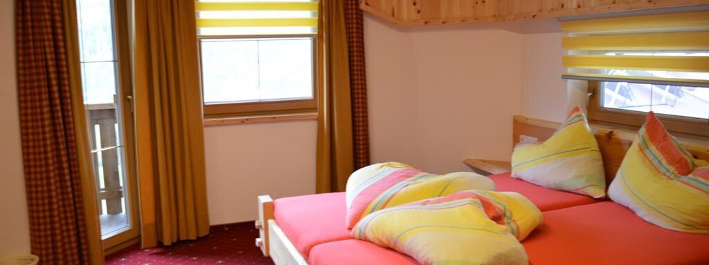A cozy bedroom with a large bed and colorful pillows. The windows are equipped with light curtains, and there is a balcony with a view. - Am Trogwohl - Sölden