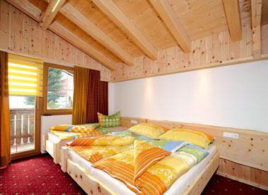 A cozy bedroom with a wooden ceiling and colorful bedcovers. There is a window with blinds and a balcony. - Am Trogwohl - Sölden