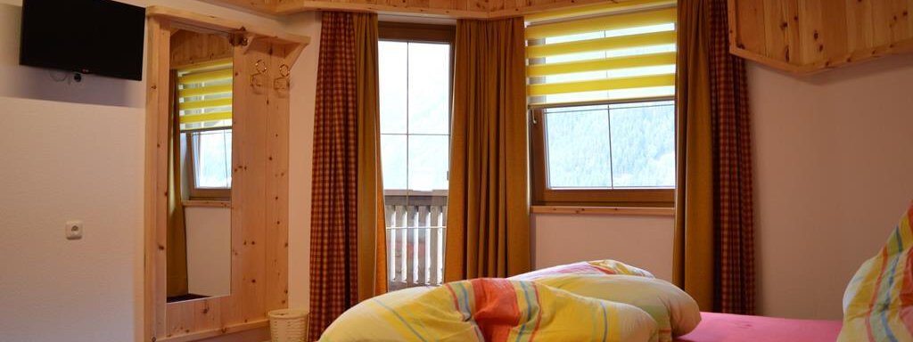 A cozy bedroom with wood paneling and large windows. The bed is equipped with colorful pillows, and a television is mounted on the wall. - Am Trogwohl - Sölden