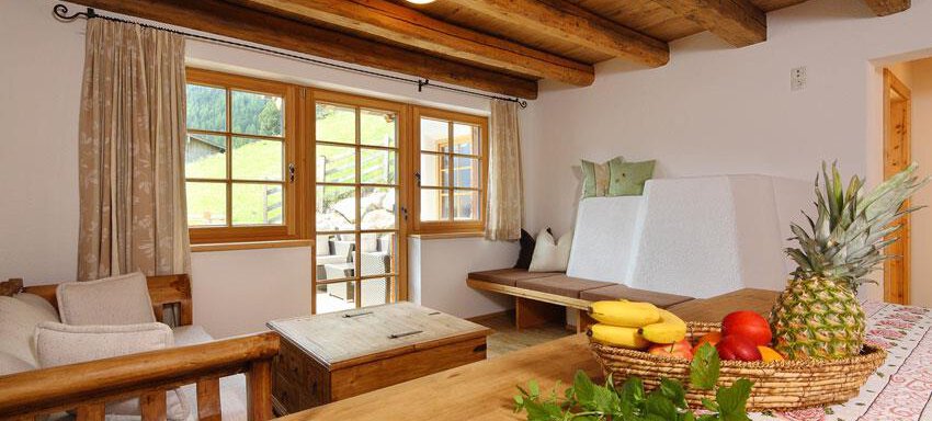 A cozy living space with a wooden ceiling and large windows. On the table sits a fruit bowl with pineapples, bananas, and apples. - Andis Almdorf - Sölden