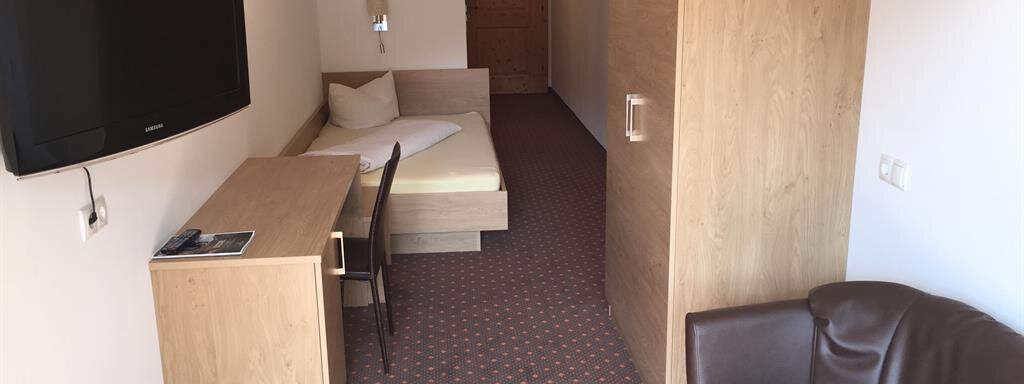 A simply furnished hotel room with a single bed, a desk, and a television. The walls are light, and there is a cozy armchair in the corner. - Andis Almdorf - Sölden