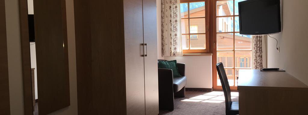 A simple, bright room with a bed, a desk, and a television. The window offers a view of a building outside. - Andis Almdorf - Sölden
