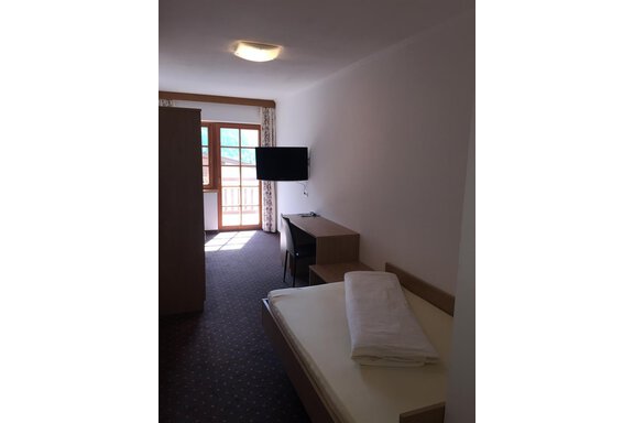 A simple hotel room with a single bed, a desk, and a television. It has a window overlooking a balcony and plenty of daylight. - Andis Almdorf - Sölden