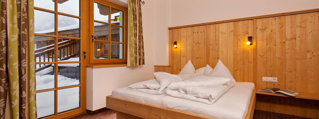 A cozy bedroom with a double bed and wooden-paneled walls. Large windows offer a view of the snow outside. - Andis Almdorf - Sölden