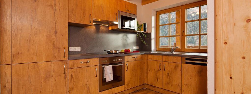 A modern kitchen with wooden cabinets and wooden flooring. It is well equipped with a stove, oven, and a large window for natural light. - Andis Almdorf - Sölden