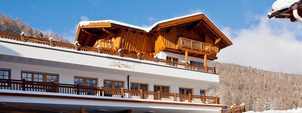 A modern building in Alpine style, surrounded by snow and fir trees. The clear blue sky provides a beautiful winter atmosphere. - Andis Almdorf - Sölden