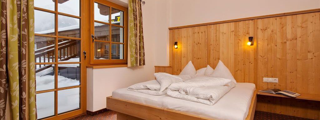 A cozy bedroom with a wooden frame bed and white beddings.  
Large windows provide plenty of daylight and a view of the snow-covered landscape. - Andis Almdorf - Sölden