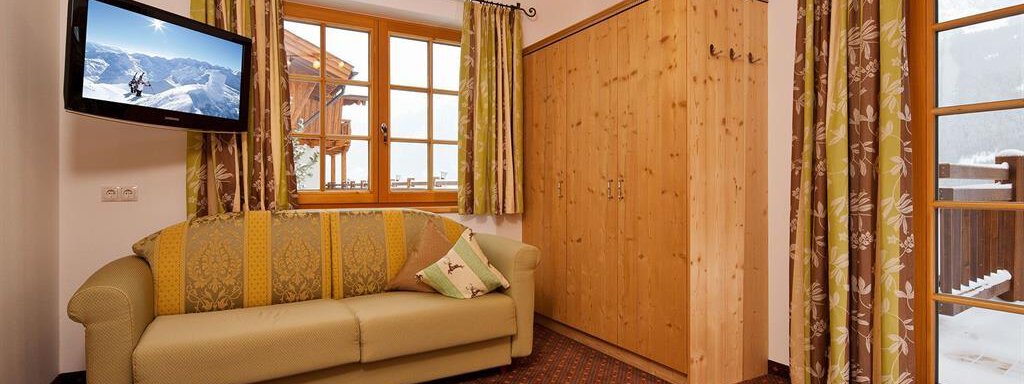 A cozy living room with a beige sofa, a television, and large windows. The curtains are green patterned, and a wooden cabinet complements the space. - Andis Almdorf - Sölden
