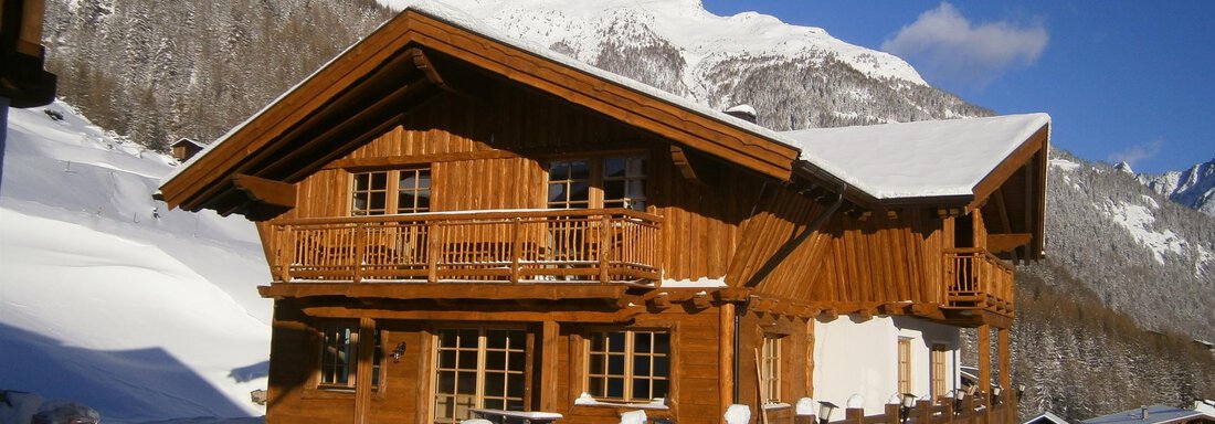 A cozy wooden house in the snow, surrounded by majestic mountains. The sky is clear and blue, creating a beautiful winter impression. - Andis Almdorf - Sölden