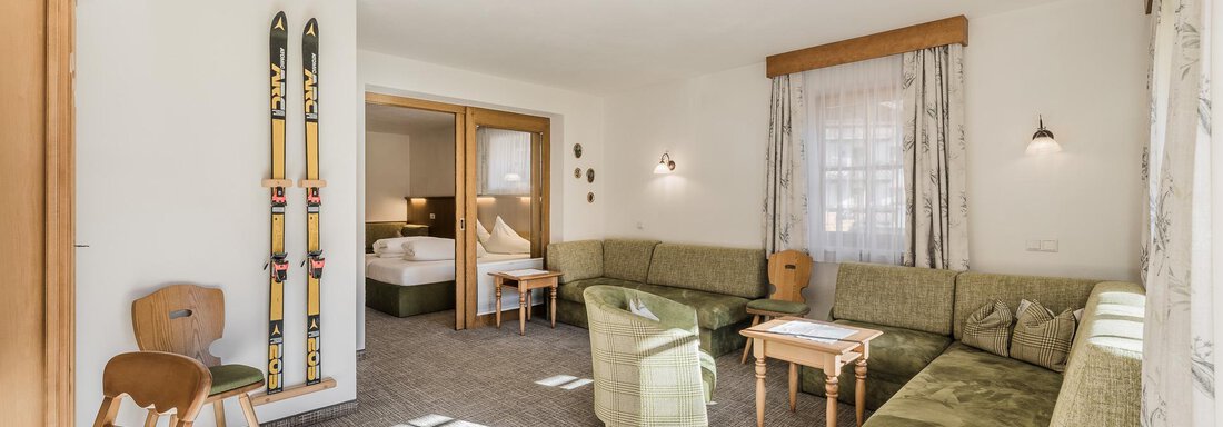 A cozy living space with a large green couch and wooden chairs. Skis hang on the wall, and there is plenty of daylight coming through the windows. - Andre Arnold - Boutique Hotel - Sölden
