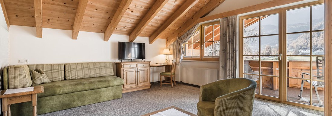 A cozy room with wooden ceilings and modern furniture. There is a sitting area, a table, and large windows with a view of the mountains. - Andre Arnold - Boutique Hotel - Sölden