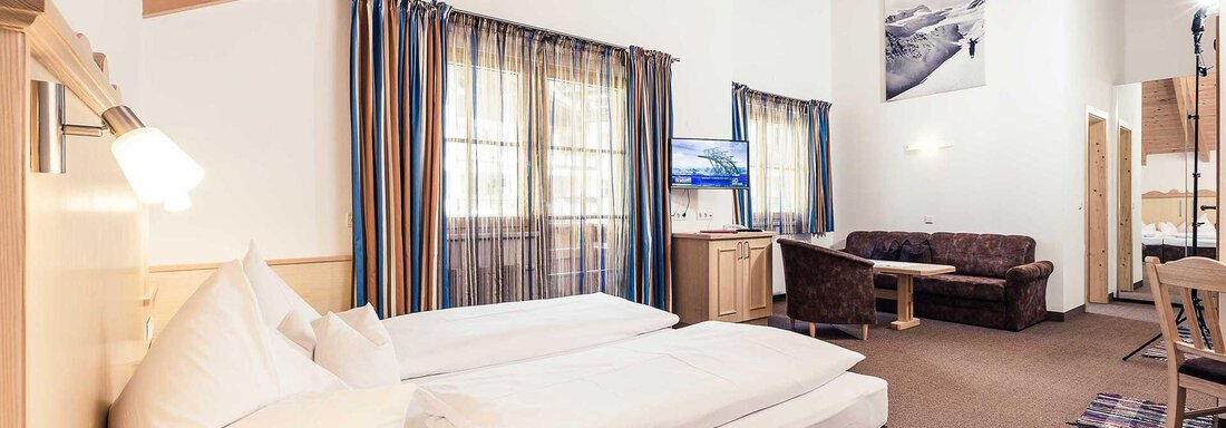 A cozy hotel room with two beds and a sitting area. Large windows let in plenty of light, and the wooden ceiling creates a warm atmosphere. - Andre Arnold - Boutique Hotel - Sölden