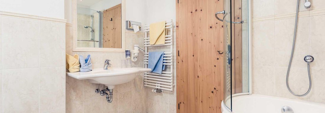 A modern bathroom with a bathtub and shower. The walls are brightly tiled, and there is a wooden shelf as well as towels in soft colors. - Andre Arnold - Boutique Hotel - Sölden