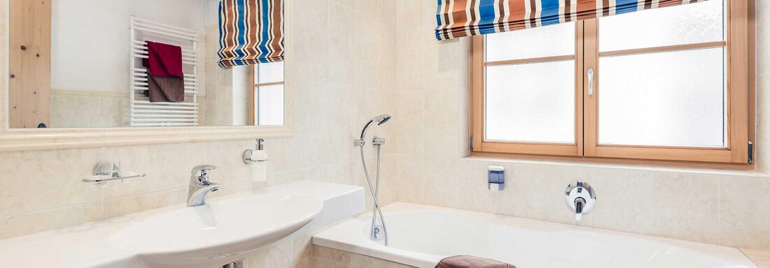 A modern bathroom with a bathtub and a sink. The windows are decorated with a striped curtain. - Andre Arnold - Boutique Hotel - Sölden