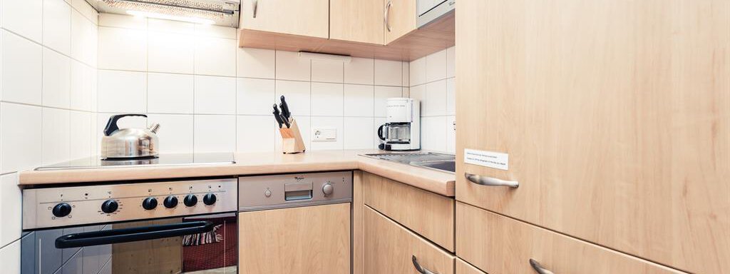 A modern kitchen with wooden cabinets and white tiles. It includes a stove, a microwave, and a kettle. - Andre Arnold - Boutique Hotel - Sölden