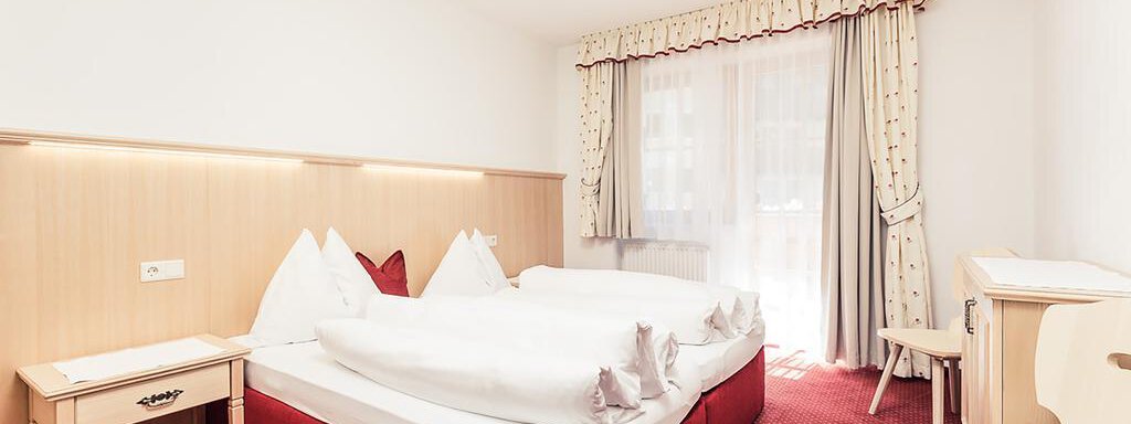 A simple, bright bedroom with two beds and red carpet. Large windows with curtains provide plenty of daylight. - Andre Arnold - Boutique Hotel - Sölden