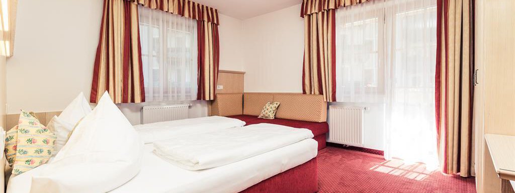 A cozy hotel room with a double bed and a seating area. Large windows let in plenty of light and the decoration is in warm colors. - Andre Arnold - Boutique Hotel - Sölden