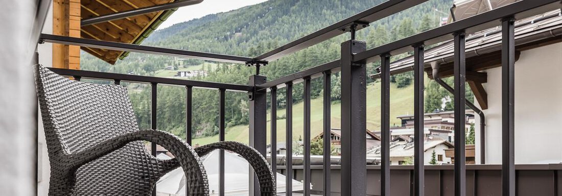 A cozy balcony with a rattan chair and a view of the mountains. The surroundings are green and sunny. - Andre Arnold - Boutique Hotel - Sölden
