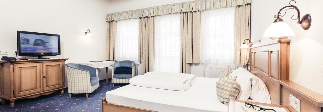 A cozy room with a woodcut bed and a television. Large windows let in plenty of light and create a pleasant atmosphere. - Andre Arnold - Boutique Hotel - Sölden